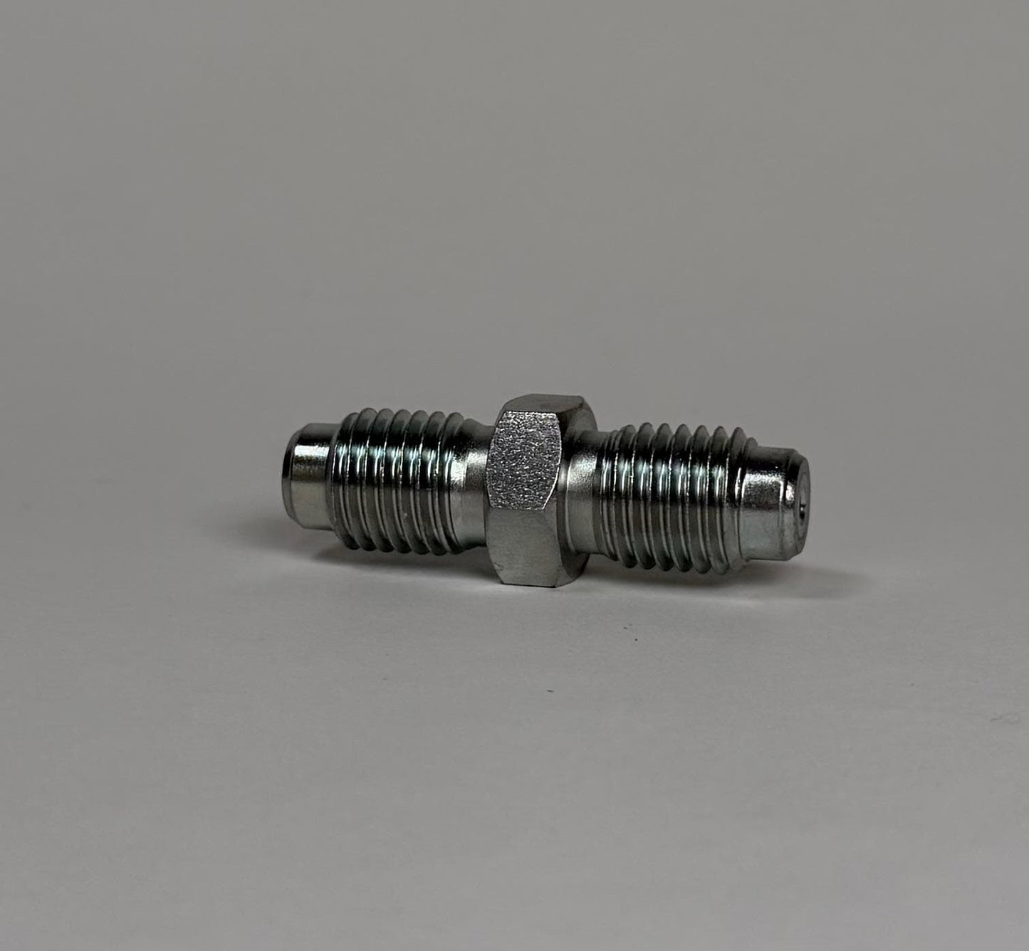 A Stauff Joiner, a metal double-ended threaded fitting with a hexagonal center, is lying horizontally on a plain gray surface.